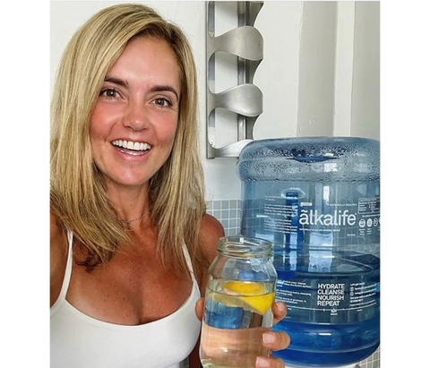a girl holding a glass of alkaline water in front of a 15L bottle of ãlkalife naturally alkaline mineral water